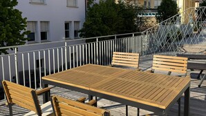 Eine Holzterrasse mit Tisch und Bänken, umgeben von einem weißen Geländer, mit Gebäuden im Hintergrund.