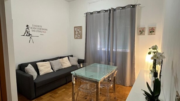 A modern living room with a glass-top dining table, a dark sofa with cushions, a white cabinet, and a potted orchid. - BalduinaView, in the residential neighborhood of Balduina just minutes from the Vatican (Roma)
