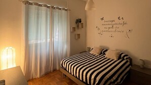 A bedroom with a bed, a bedside table, a lamp, a wall decoration, and a window with curtains. - BalduinaView, in the residential neighborhood of Balduina just minutes from the Vatican (Roma)
