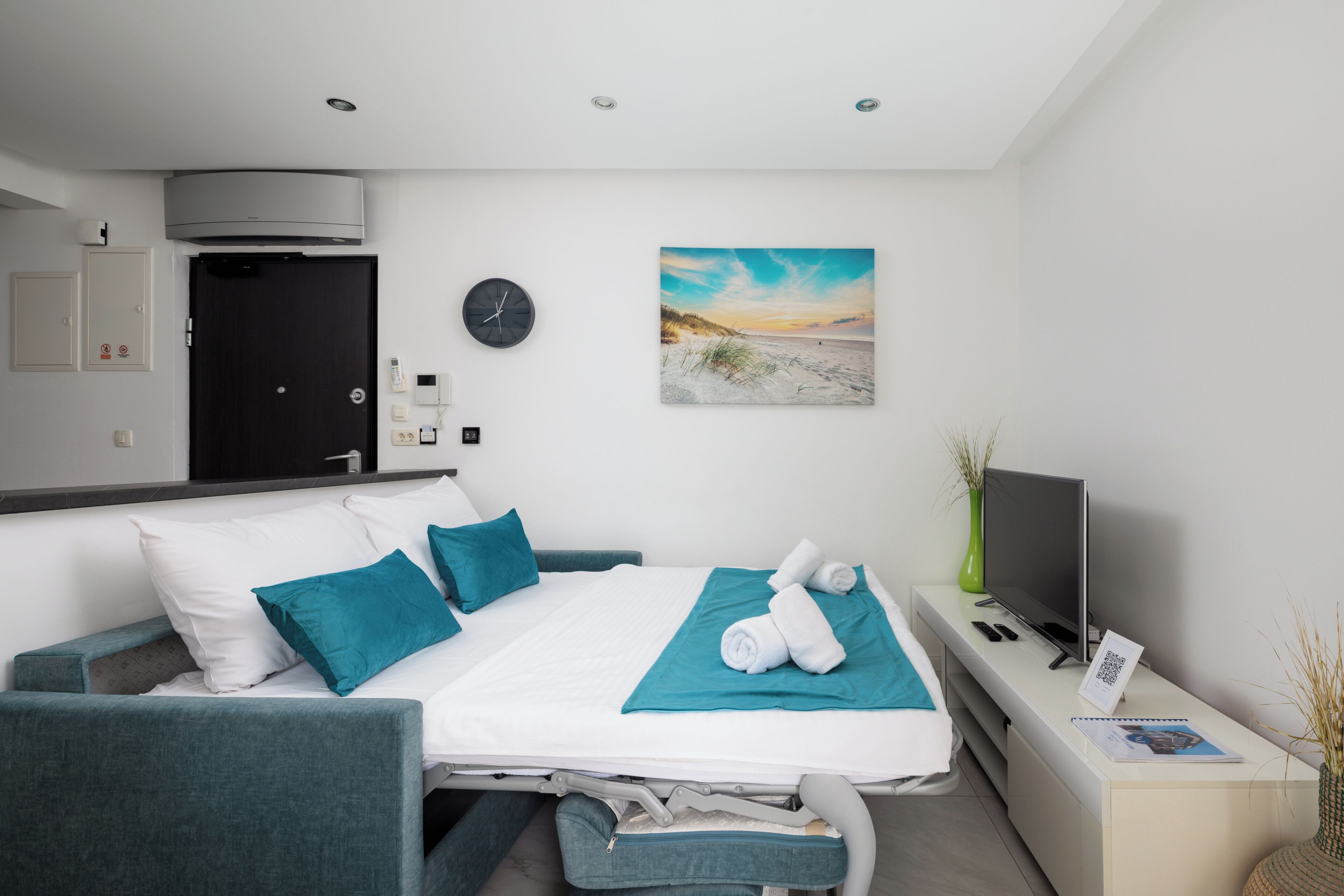 A hotel room with a bed, a TV, a clock, and a painting on the wall.