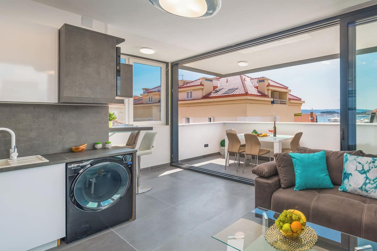 A modern apartment with a kitchen, a washing machine, a dining area, and a sofa with a view of the city.