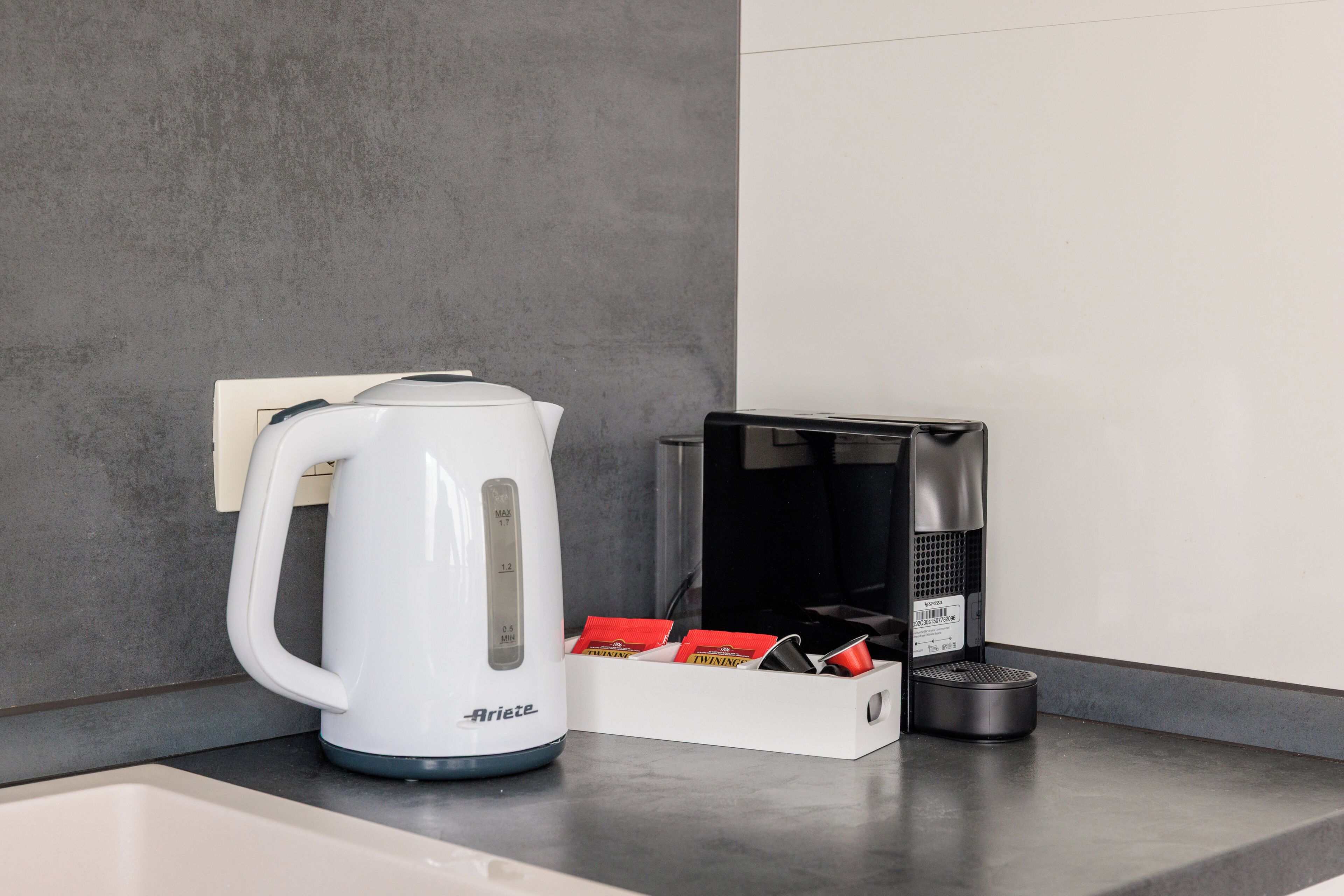 Una tetera eléctrica blanca con temporizador, una caja de bolsitas de té y una cafetera negra sobre una encimera.