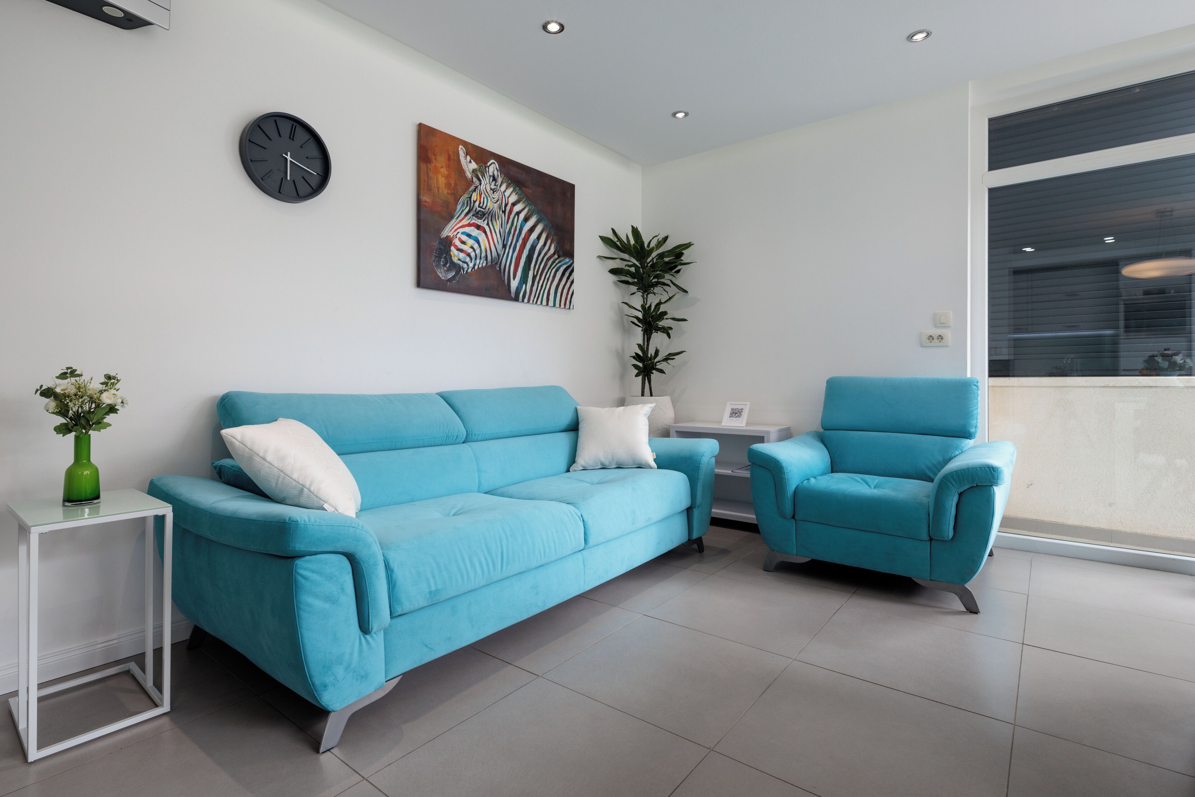 A modern living room with a blue sofa, a matching armchair, a clock, a zebra painting, and a potted plant.