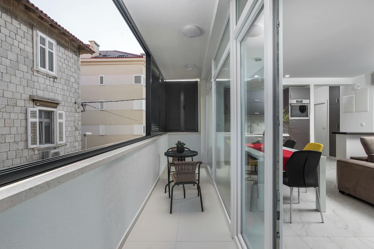 A narrow balcony with a table and chairs, a sliding glass door leading to a kitchen, and a view of neighboring buildings.