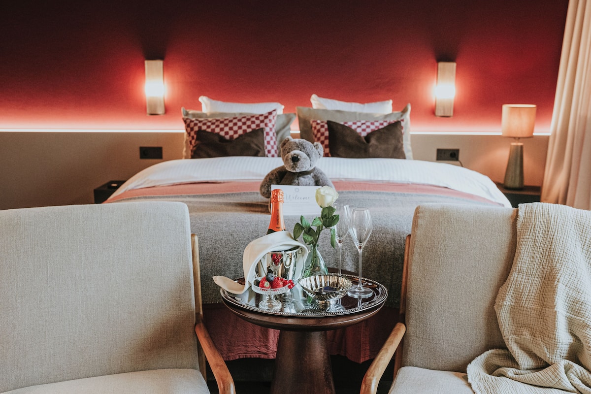 Habitación de hotel con una cama bien hecha, un osito de peluche sobre la cama, y una bandeja con una botella, vasos y un cuenco de fruta.