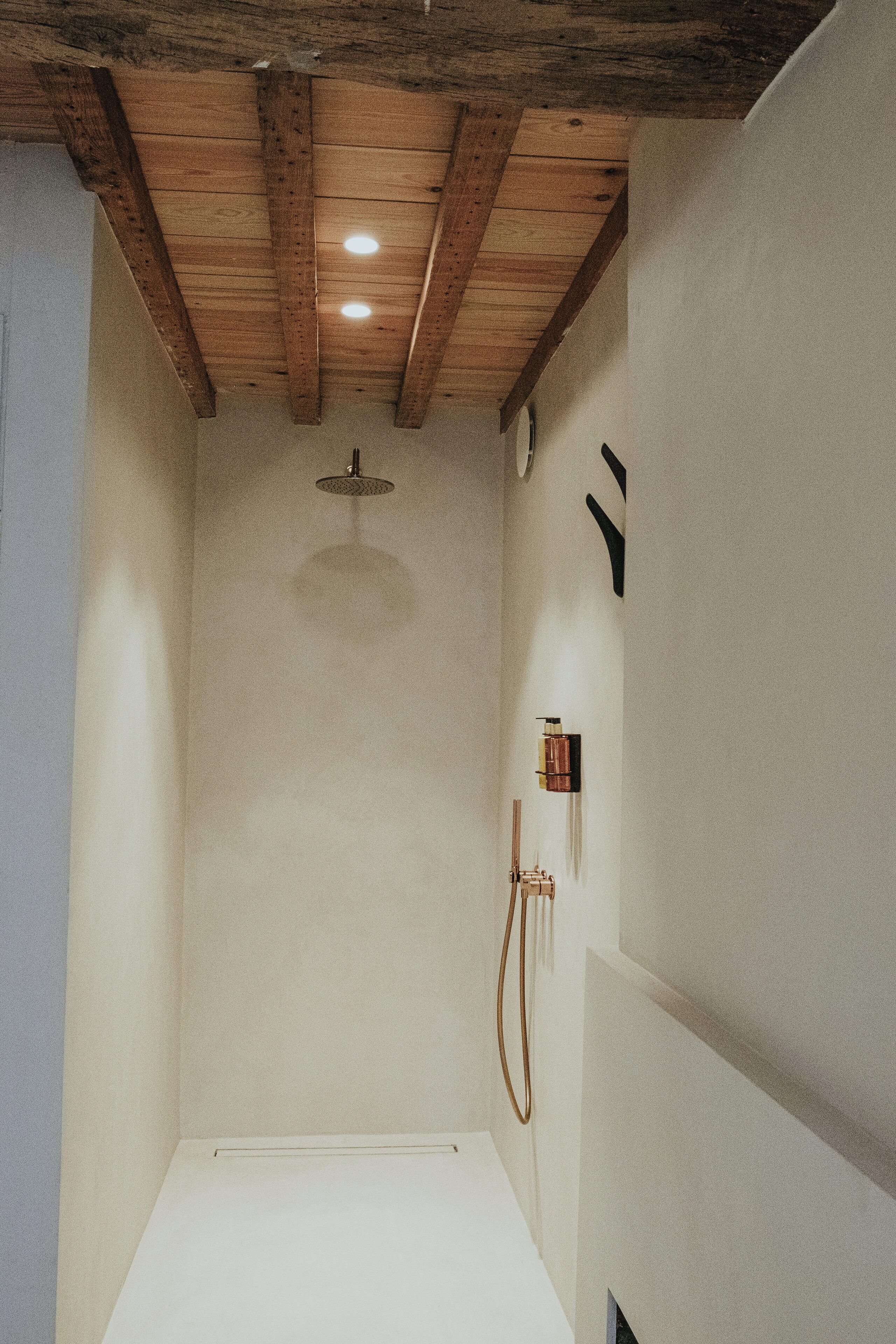 A narrow, white-walled shower with wooden beams on the ceiling and a single light fixture.