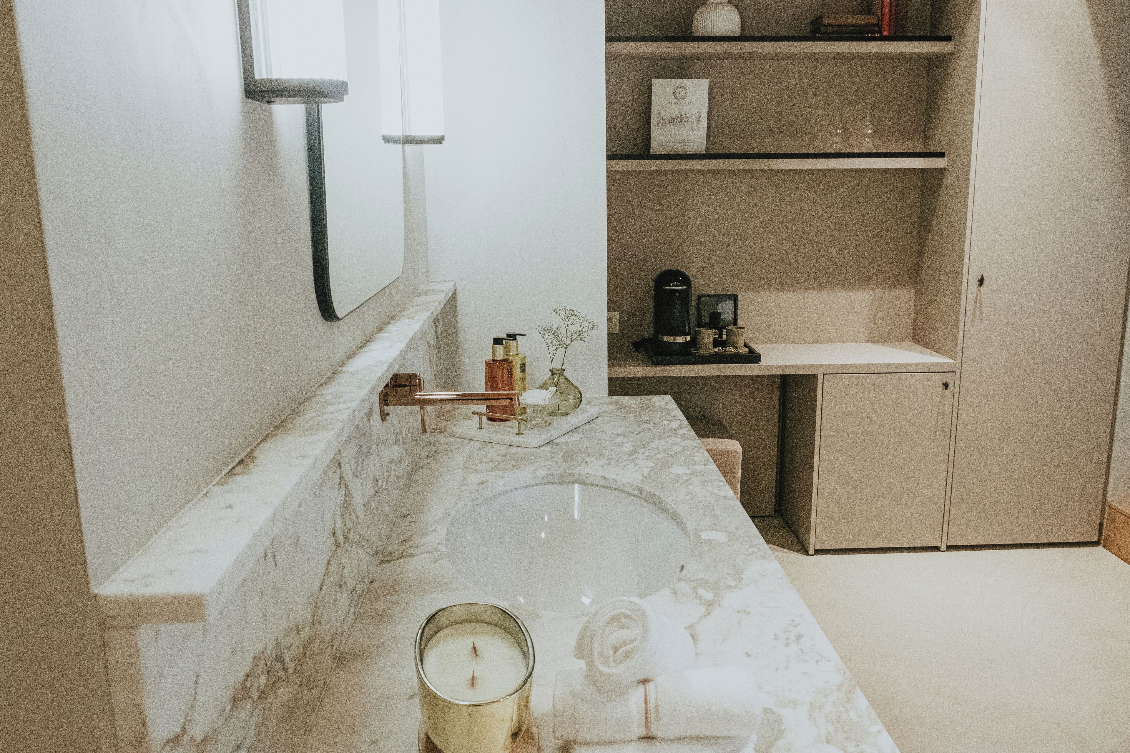 A bathroom with a marble sink, a mirror, a shelf with toiletries, and a cabinet.