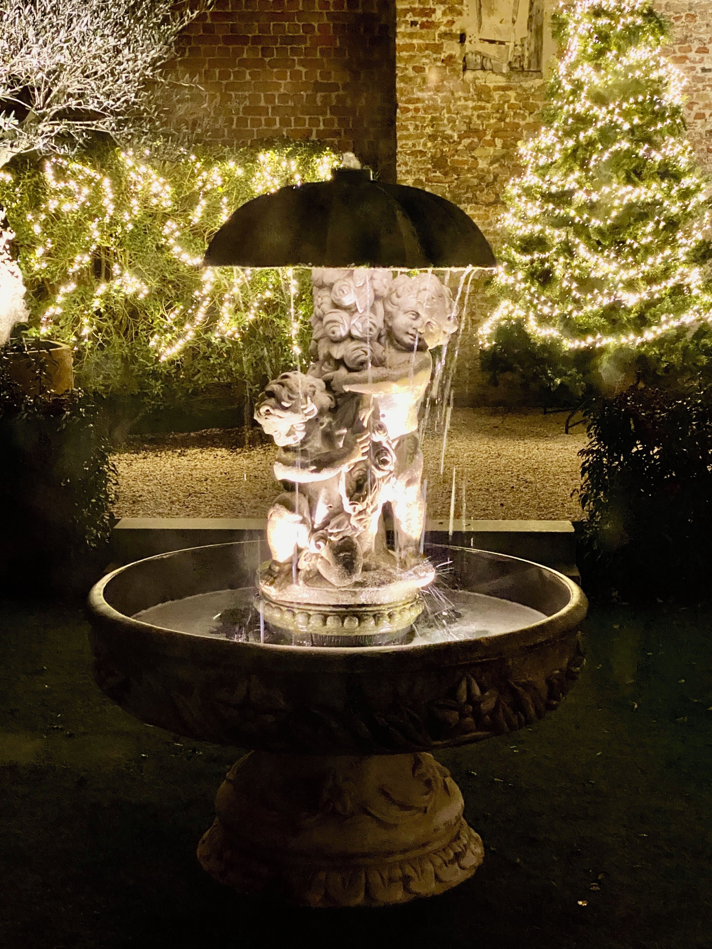 A fountain with a decorative sculpture, surrounded by greenery and illuminated lights.