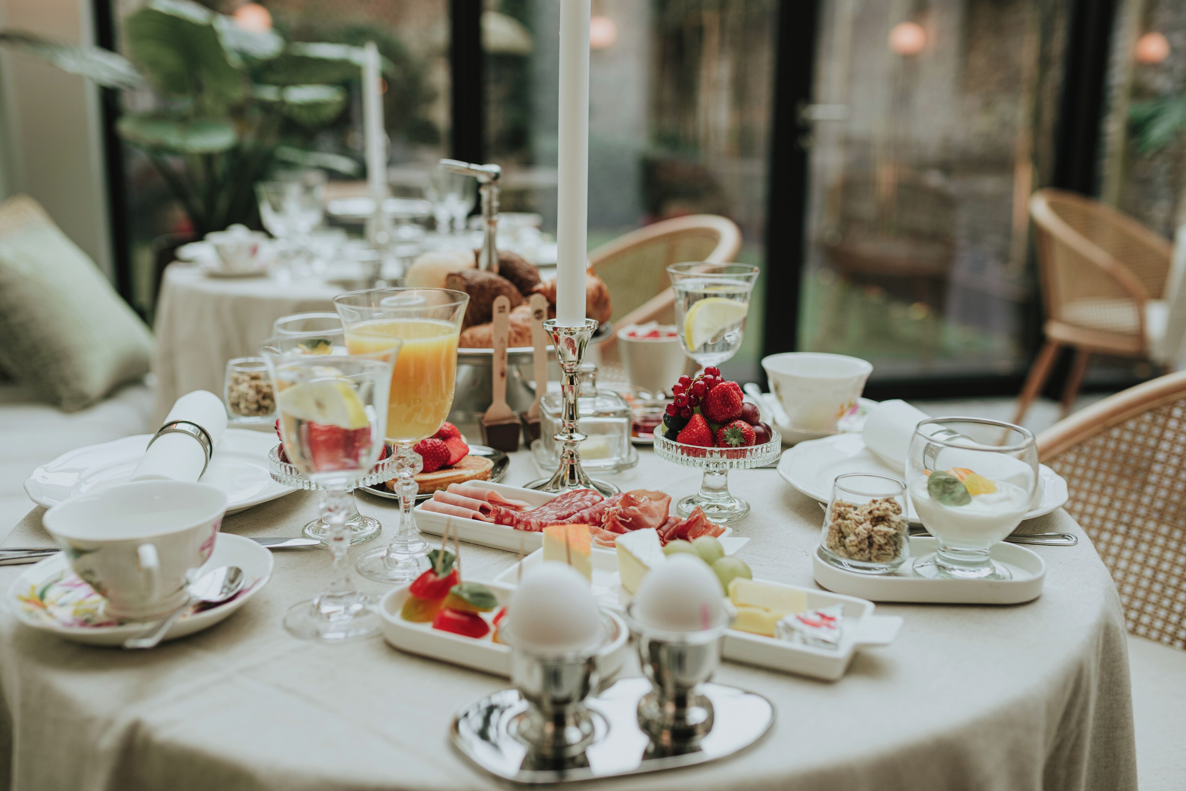 A breakfast table set with various food items, including fruits, cheeses, and beverages, in a well-lit room with a view of greenery outside.