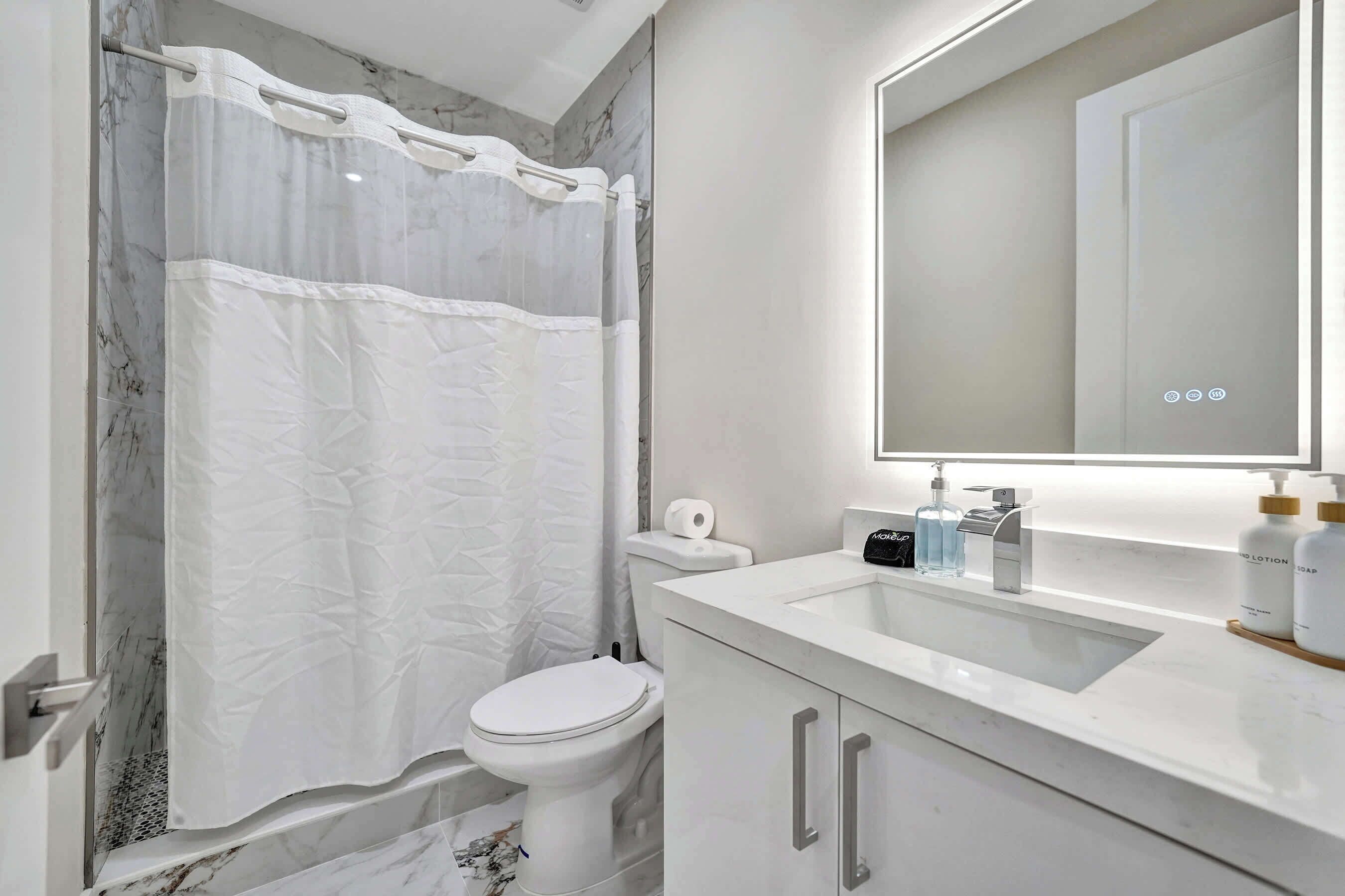 A bathroom with a white toilet, a shower curtain, a sink with a mirror, and toiletries on the counter.