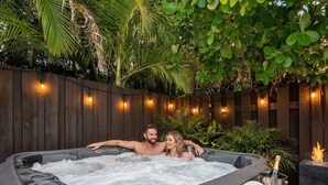 A hot tub with two people, surrounded by a wooden fence and string lights, with a bottle of champagne and a towel on a table nearby. - 6 BR Villa Steps to Ocean, Pool, Sauna & Jacuzzi (Fort Lauderdale)