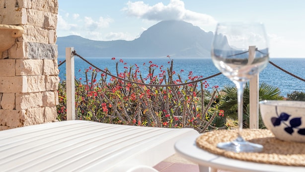 Una terraza con una mesa, una copa de vino y un cuenco, con vistas al mar, flores y una montaña.