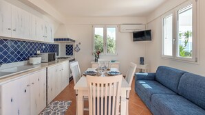 Cocina compacta con armarios blancos, salpicadero de azulejos azules y comedor con mesa y sillas blancas. Un sofá azul está ubicado cerca de una ventana con un televisor montado encima.