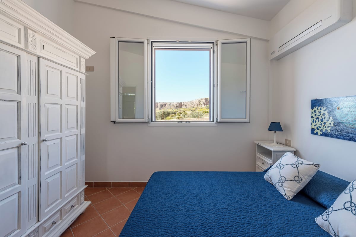 A bedroom with a bed, a wardrobe, a window with a view, a nightstand, and a lamp.