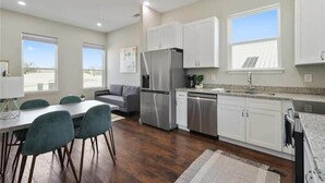 Una cocina moderna con armarios blancos, electrodomésticos de acero inoxidable y un comedor con sofá gris y sillas color turquesa.