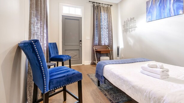 Ein Hotelzimmer mit einem Bett, zwei blauen Polsterstühlen, einem kleinen Tisch, einem Wandbild und einer Tür mit Fenster.