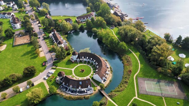 An aerial view of a lakeside property with a circular building, a tennis court, and a dock with boats. - 'Pepperpot' Lodge on Isis & Windrush Lakes with Great On-site Facilities (South Cerney)