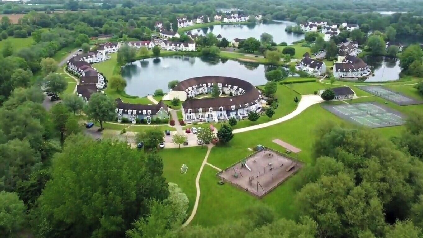 An aerial view of a residential area with houses arranged around a central lake, a tennis court, and a playground.