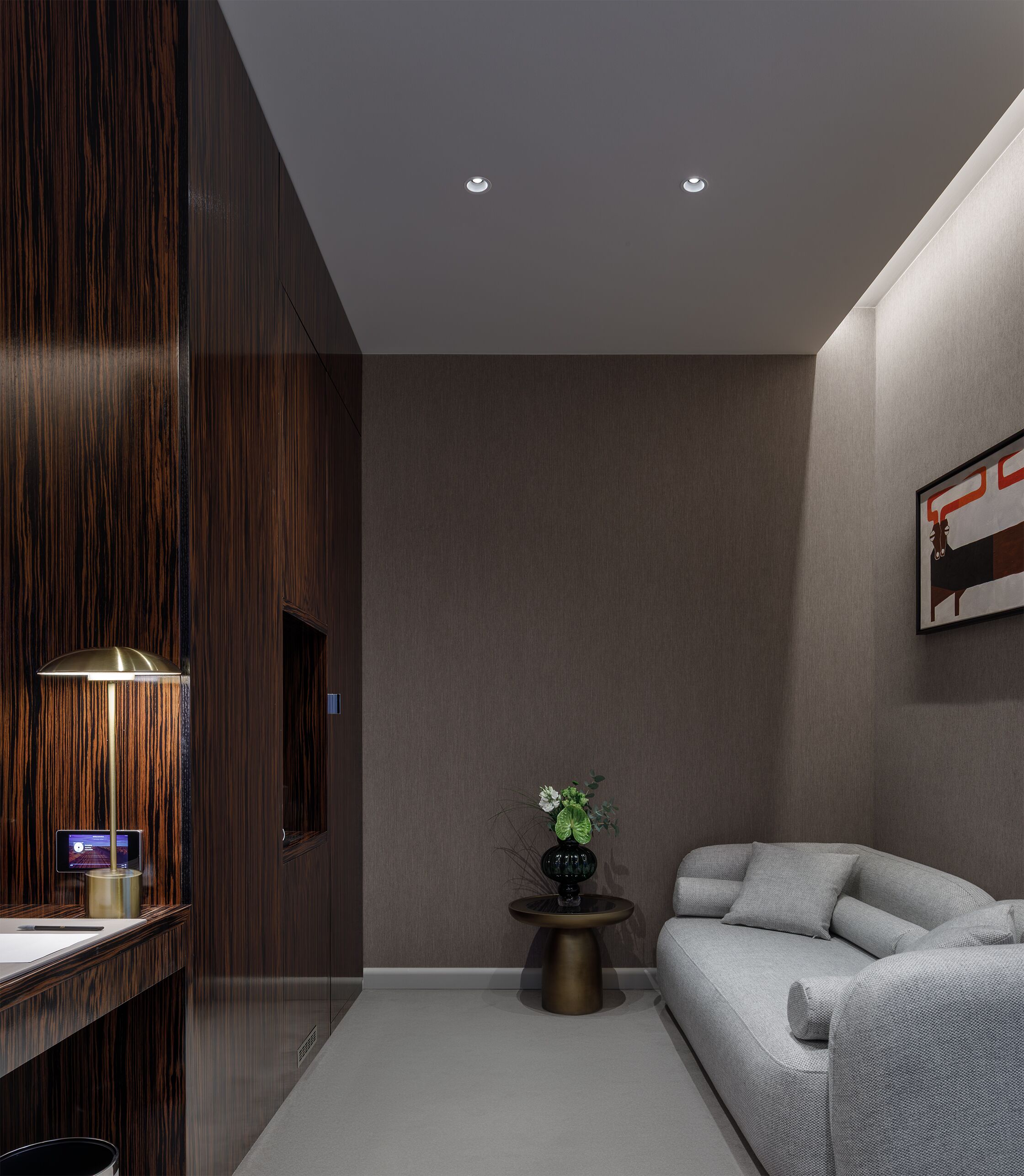 A modern hotel room with a wooden accent wall, a round side table with a vase of flowers, a grey sofa, and recessed ceiling lighting.