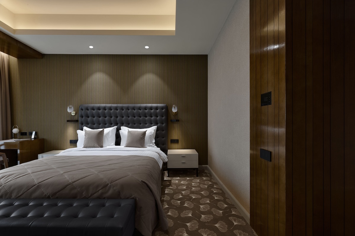 A modern hotel room with a large bed, a bedside table, a nightstand, and a wooden wardrobe.