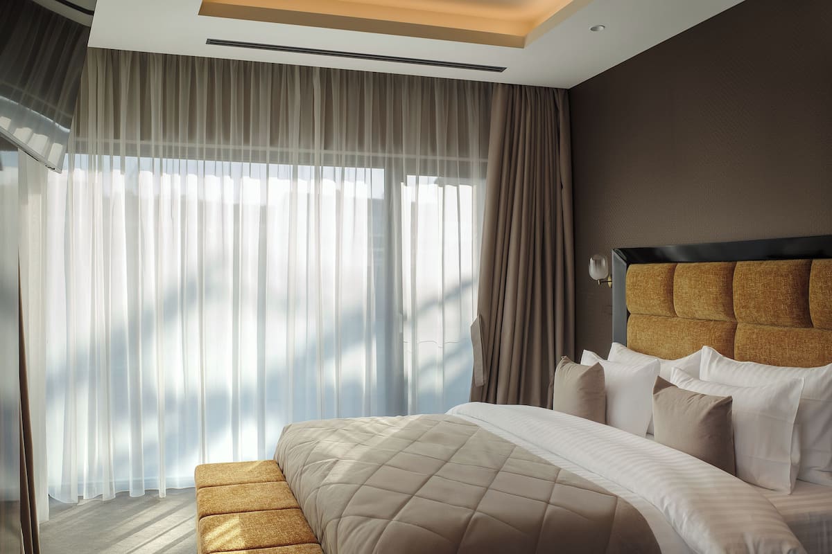 Ein modernes Hotelzimmer mit einem großen Bett, einem gepolsterten Kopfteil und transparenten Vorhängen, die natürliches Licht hereinlassen.