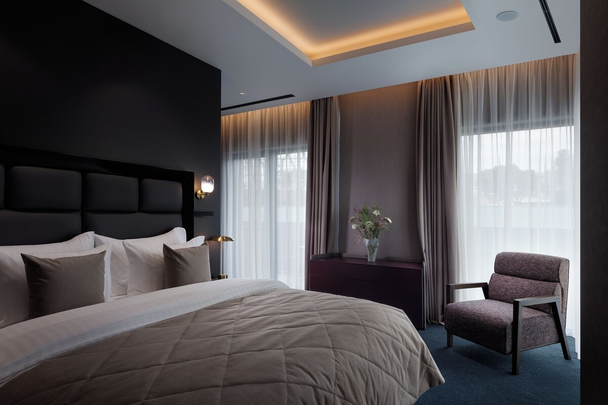 A modern hotel room with a large bed, a chair, and a window with curtains.