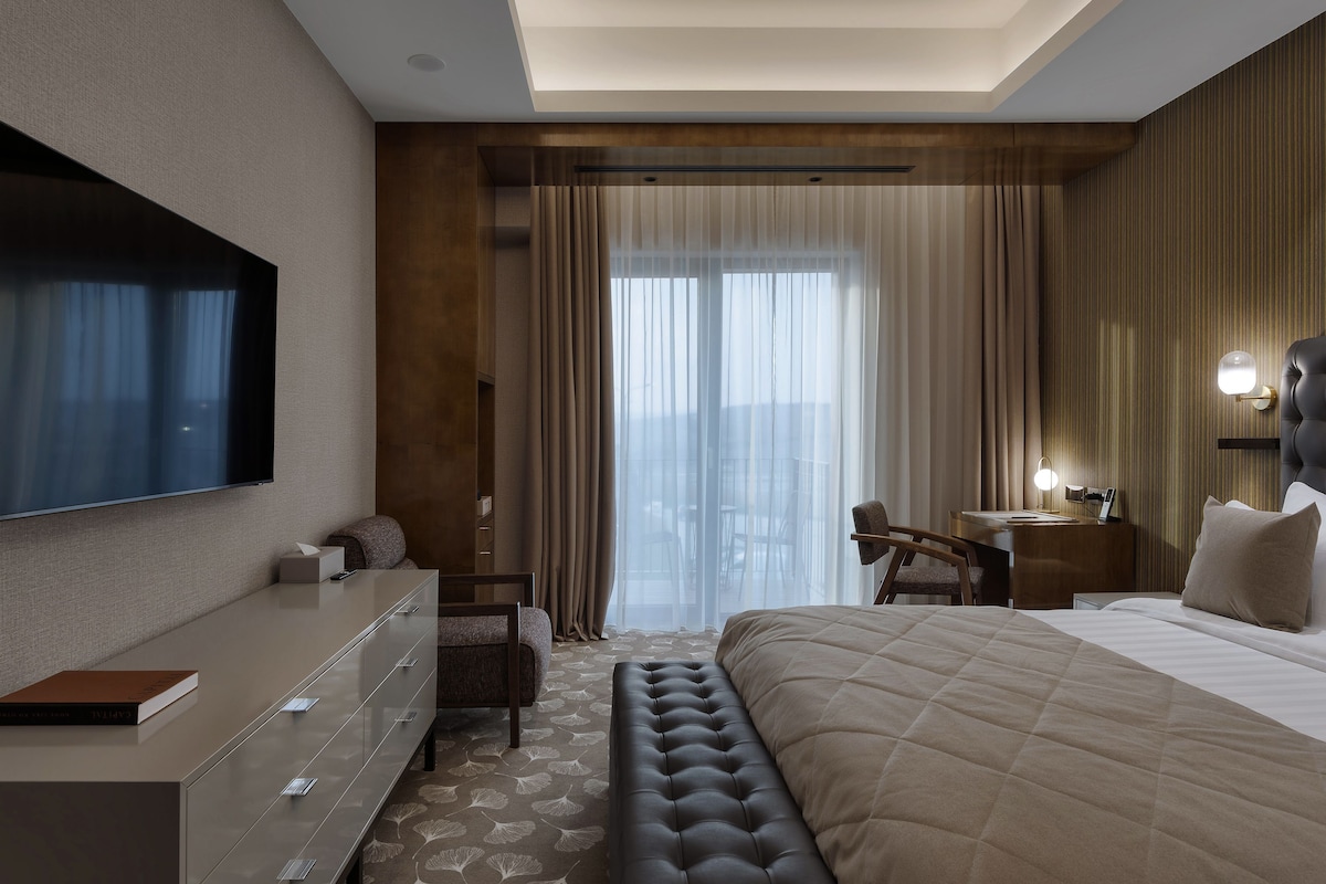 Una habitación de hotel moderna con una cama grande, un televisor de pantalla plana, un escritorio con lámpara y vistas a la ciudad a través de amplias ventanas.