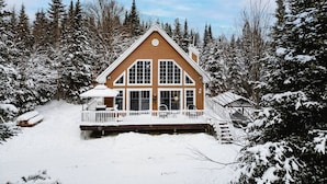 Una cabaña de troncos grande de dos pisos, con un porche envolvente y múltiples ventanas, rodeada de árboles cubiertos de nieve.