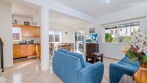 A spacious living area with a blue sofa set, a dining table, and a kitchen area with wooden cabinets and a microwave. - Villa Kos #7 - Charming Paralimni Getaway (Protaras)