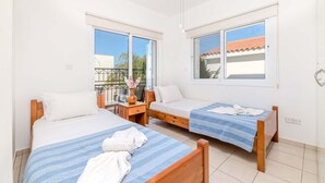 A hotel room with two single beds, a small table with a vase, and a window with a view of the outside. - Villa Kos #7 - Charming Paralimni Getaway (Protaras)