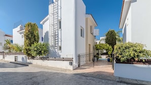 A white building with a metal staircase, surrounded by greenery and a paved street. - Villa Kos #7 - Charming Paralimni Getaway (Protaras)