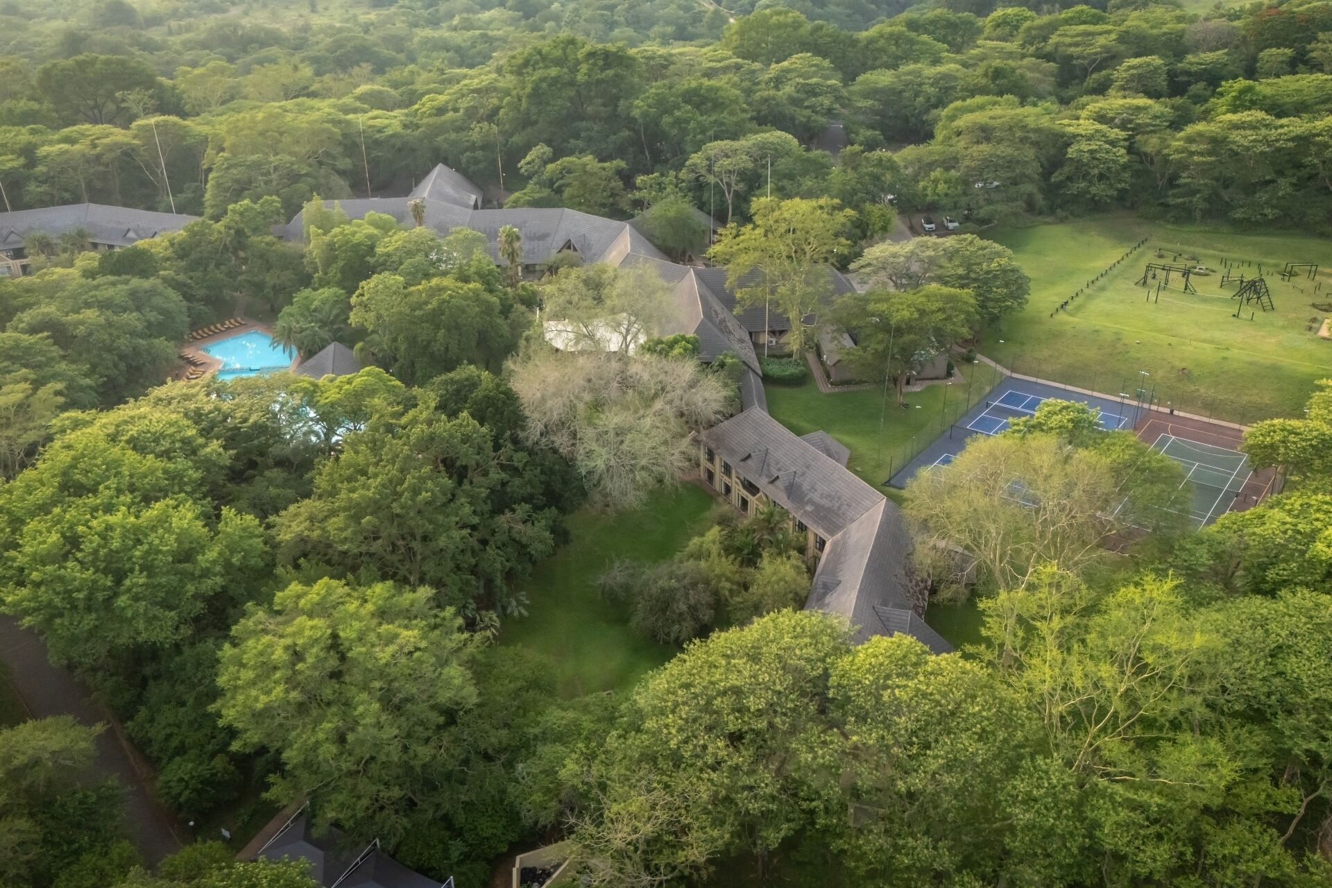 An aerial view of a resort with a swimming pool, tennis courts, and lush greenery.