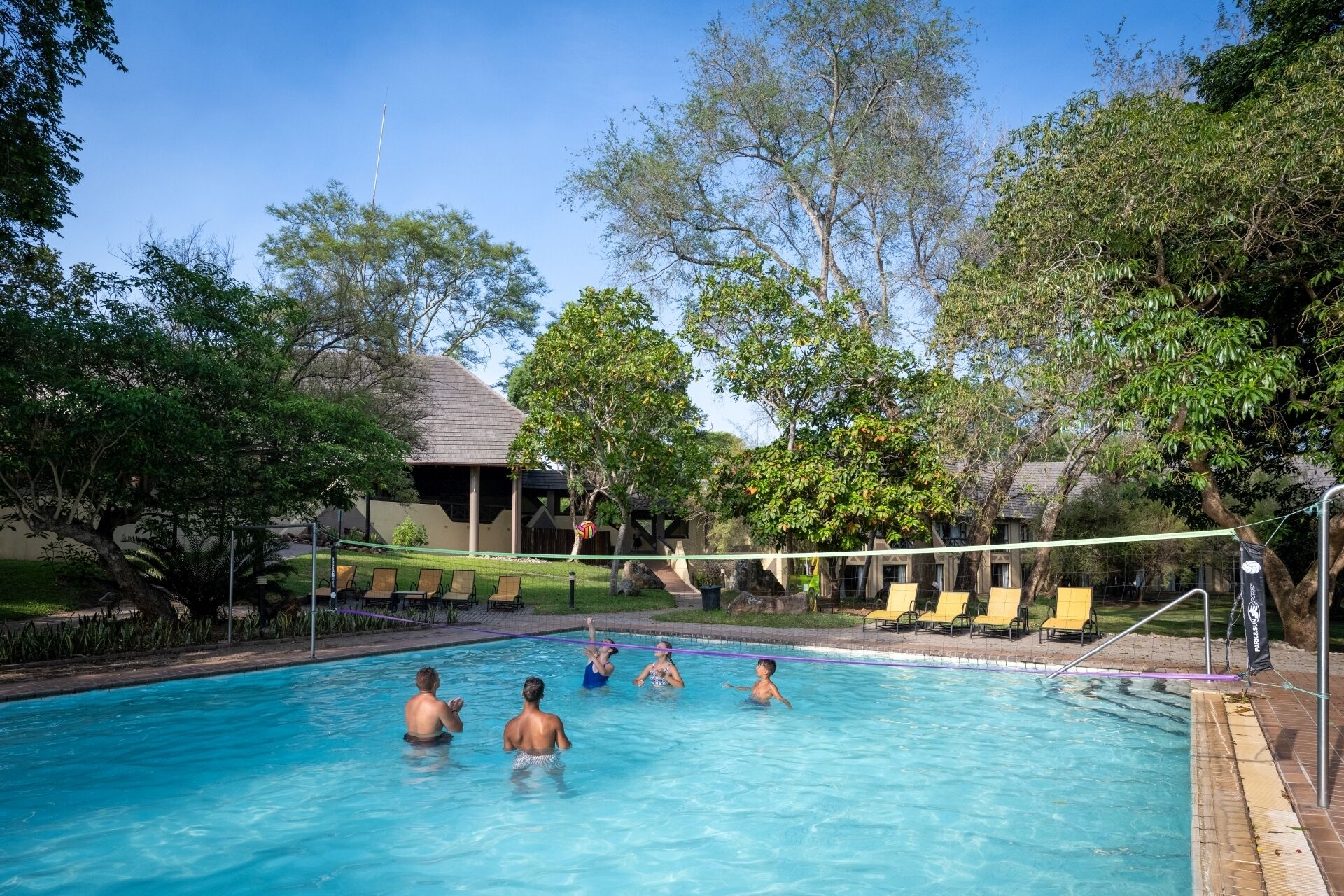 A swimming pool with people playing volleyball, surrounded by trees and a building with a sloped roof.