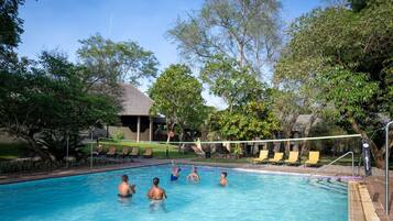 A swimming pool with people playing volleyball, surrounded by trees and a building with a sloped roof.