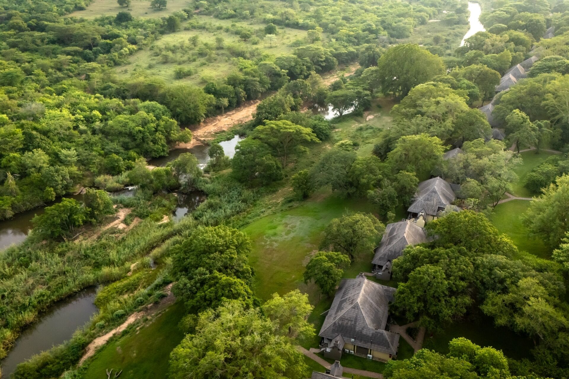 Vue aérienne d’une zone rurale avec une rivière, une végétation luxuriante et plusieurs maisons traditionnelles à toits de chaume.