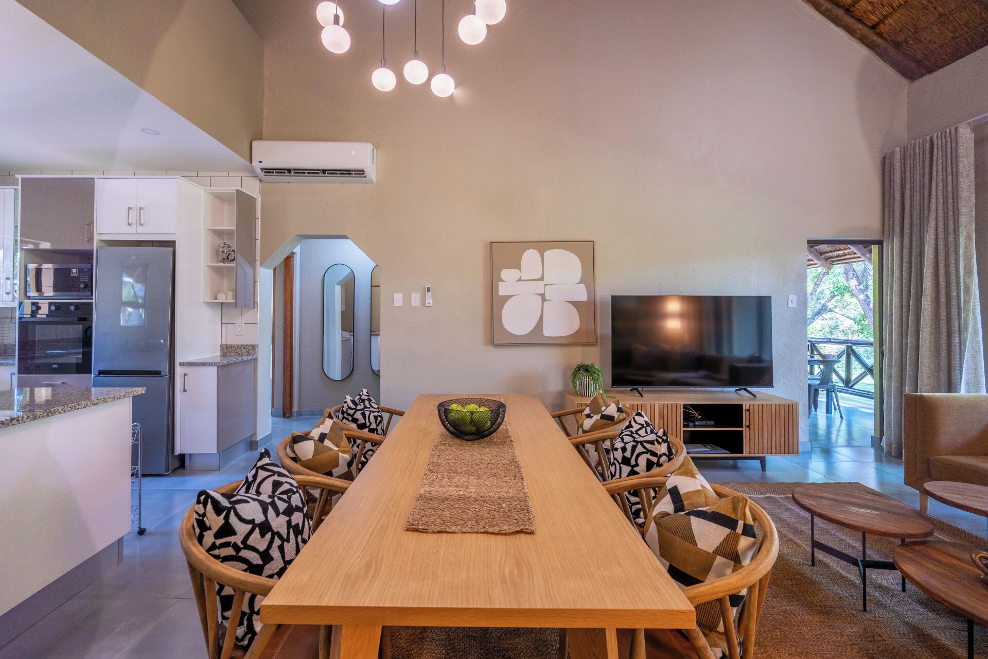 A modern kitchen with a built-in oven and refrigerator, a dining area with a wooden table and chairs, a flat-screen TV, and a view of the outdoors.