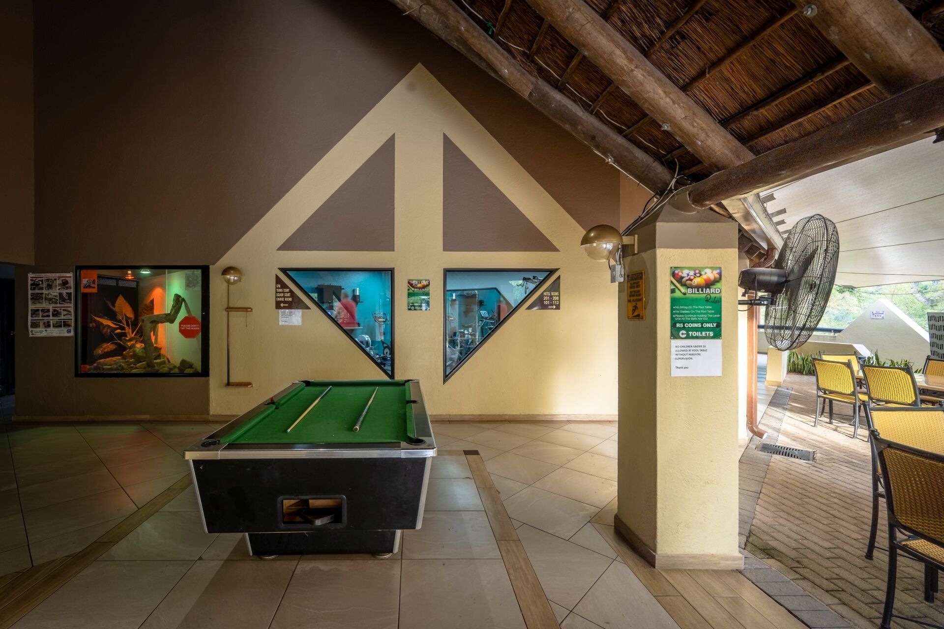 Uma sala de bilhar com mesa de bilhar, ventilador de teto e um aviso com as regras do jogo e instruções para uso de moedas.