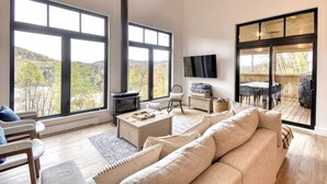 Ein geräumiges Wohnzimmer mit großen Fenstern, einem Kamin, einer Couch und einem Fernseher.