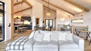 Ein geräumiger Wohnbereich mit einem grauen Ecksofa, Holzbalken an der Decke und einer Küche im Hintergrund.