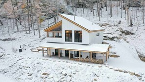 Eine moderne, zweistöckige Holzhütte mit einer großen Veranda, umgeben von einem verschneiten Wald.