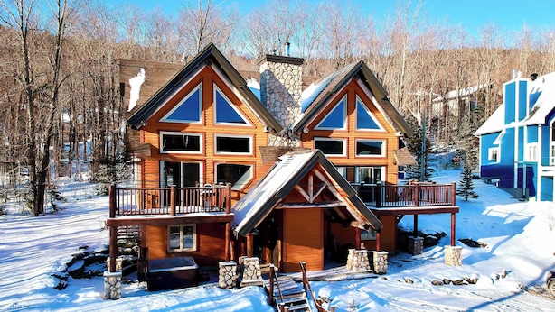 Une cabane en bois dotée d’un balcon, construite sur des fondations en pierre, et entourée d’un paysage enneigé.