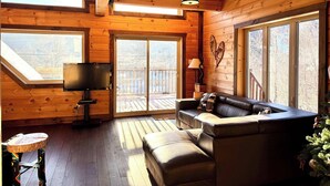 L’intérieur chaleureux d’un chalet, avec un décor en bois, un canapé en cuir, un téléviseur et de grandes fenêtres donnant sur le paysage extérieur.