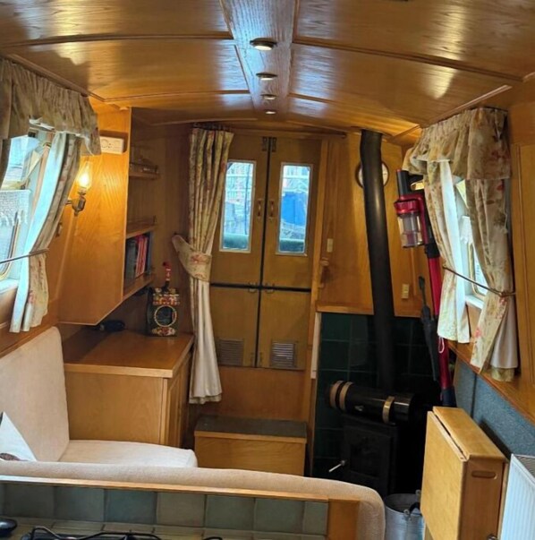 A compact RV interior with a bed, a small table with a lamp, a window with curtains, and a fireplace.