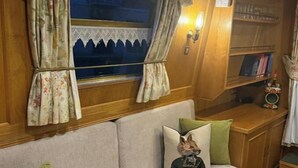 A cozy interior with a sofa, a decorative pillow, a window with curtains, and a bookshelf. - Narrowboat in Birmingham City (Birmingham)
