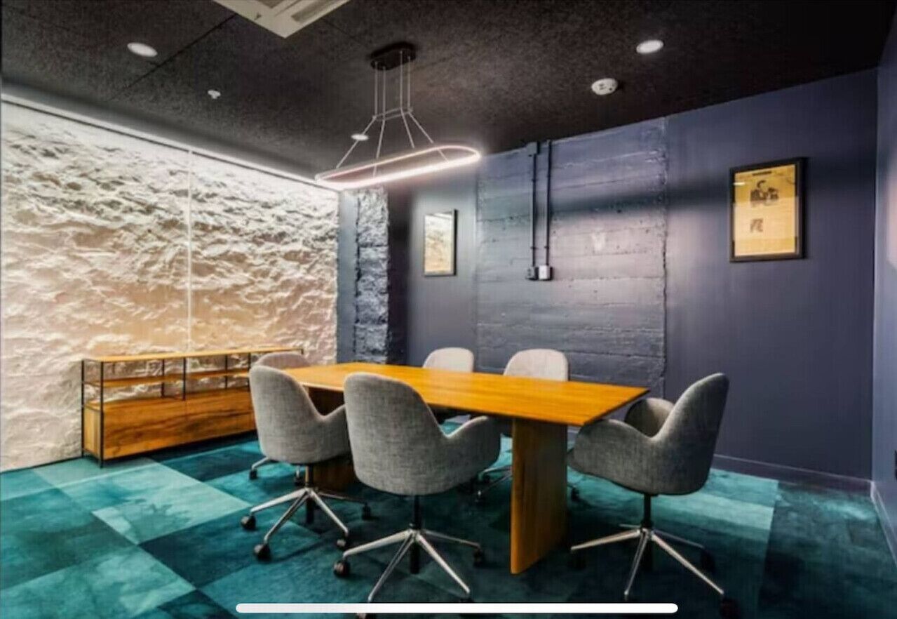 Een vergaderruimte met een houten tafel en grijze stoelen, een stenen muur en een blauw vloerkleed.