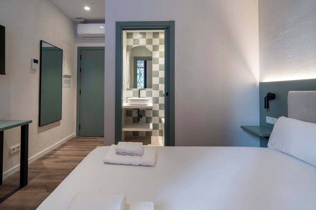A hotel room with a bed, a desk, a bathroom with a checkered tile wall, and a window.