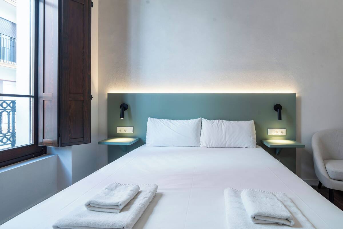 A neatly made bed with white linens and two folded towels, a nightstand with a lamp, and a chair in a room with a window and a wall-mounted light fixture.