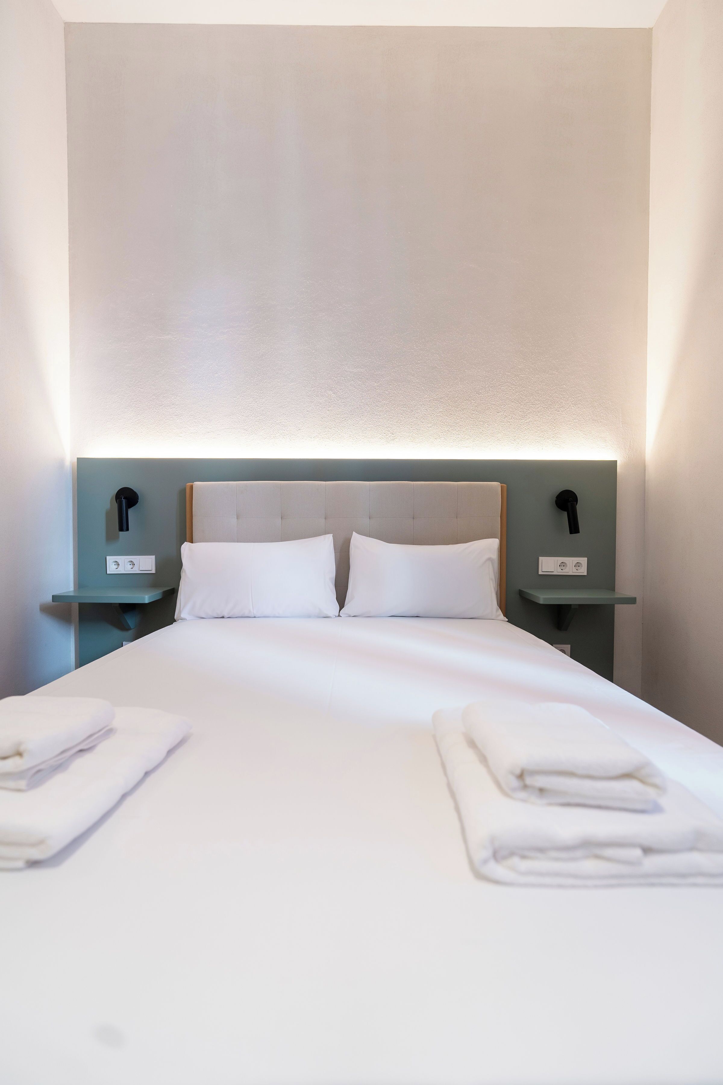 Ein ordentlich bezogenes Bett mit weißen Leinen und Kissen, zwei Nachttische mit Lampen und eine helle Wand.