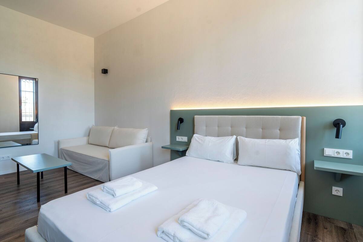 A hotel room with a large bed, a bedside table, a chair, a small table, and a mirror.