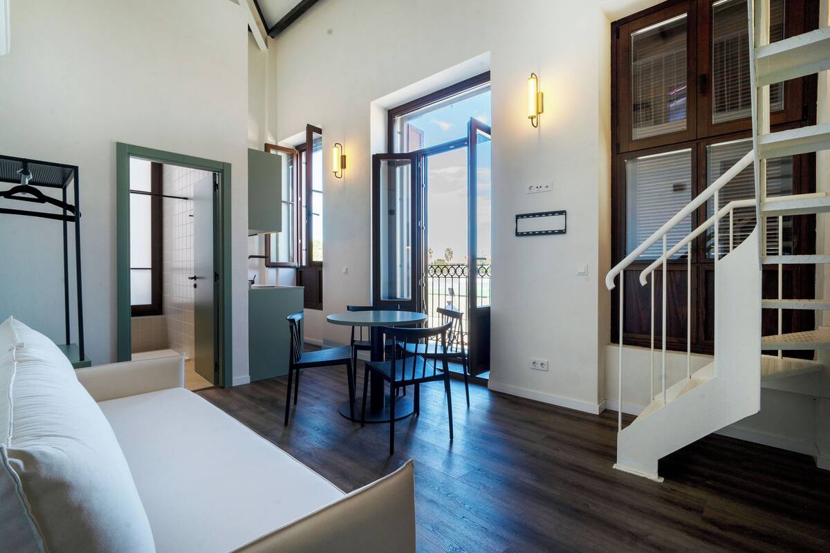 A modern hotel room with a bed, a dining area with a table and chairs, a staircase, and a view of the outdoors.